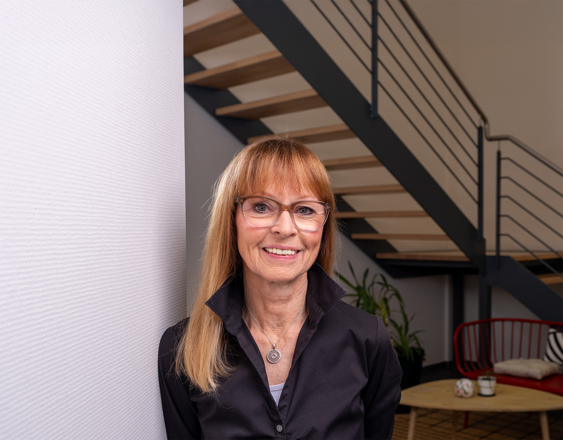 Frau mit langen blonden Haaren und Brille in einem modernen Büro mit Treppe und Pflanzen – Teil des Teams von Peter Kröner GmbH, einem familiengeführten Stahlunternehmen für die Partikelschaumindustrie.