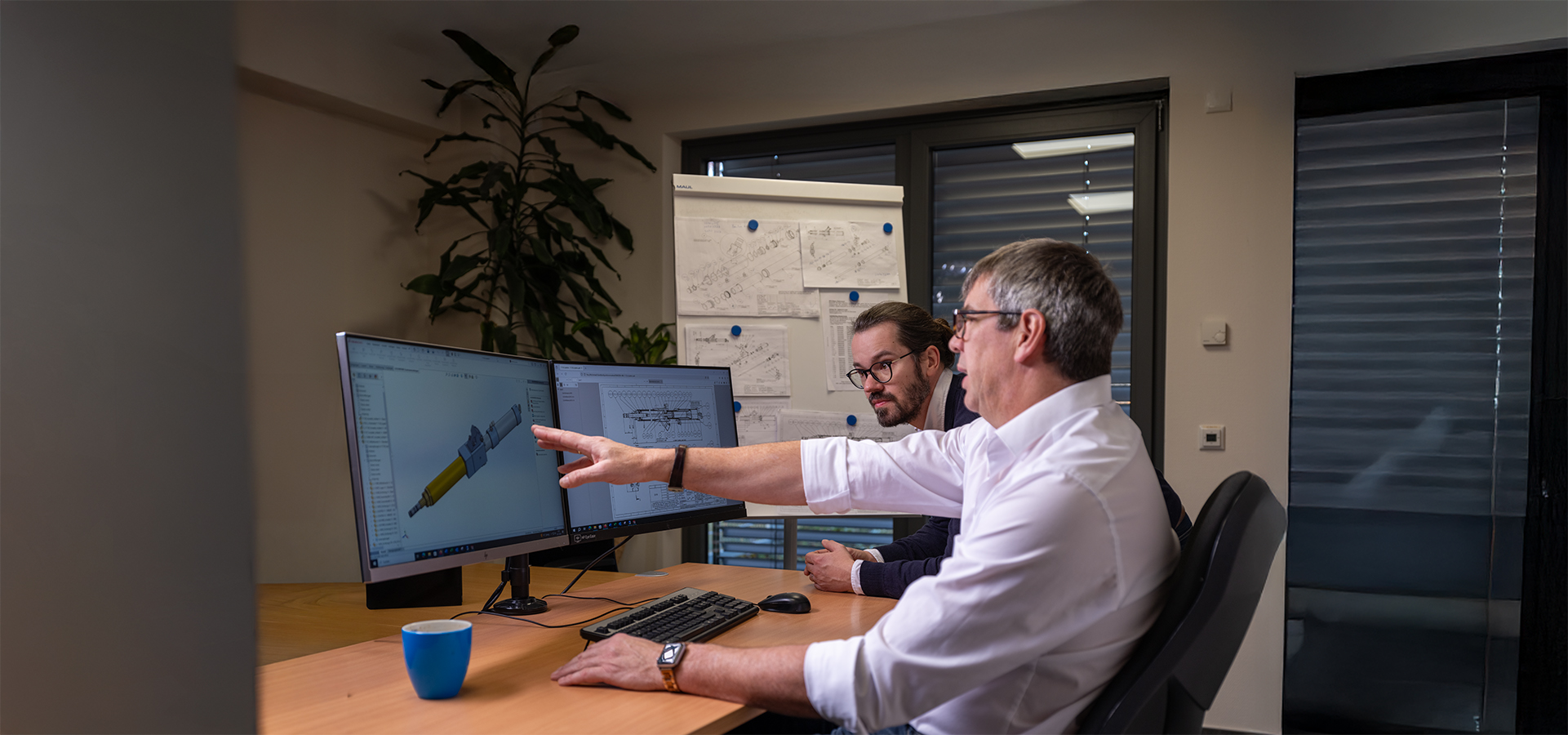 Zwei Mitarbeiter der Peter Kröner GmbH besprechen ein 3D-Modell eines Injektors am Computer, im Hintergrund technische Zeichnungen – das Unternehmen entwickelt und fertigt Lösungen für die Partikelschaumindustrie wie Injektoren und Zubehör.