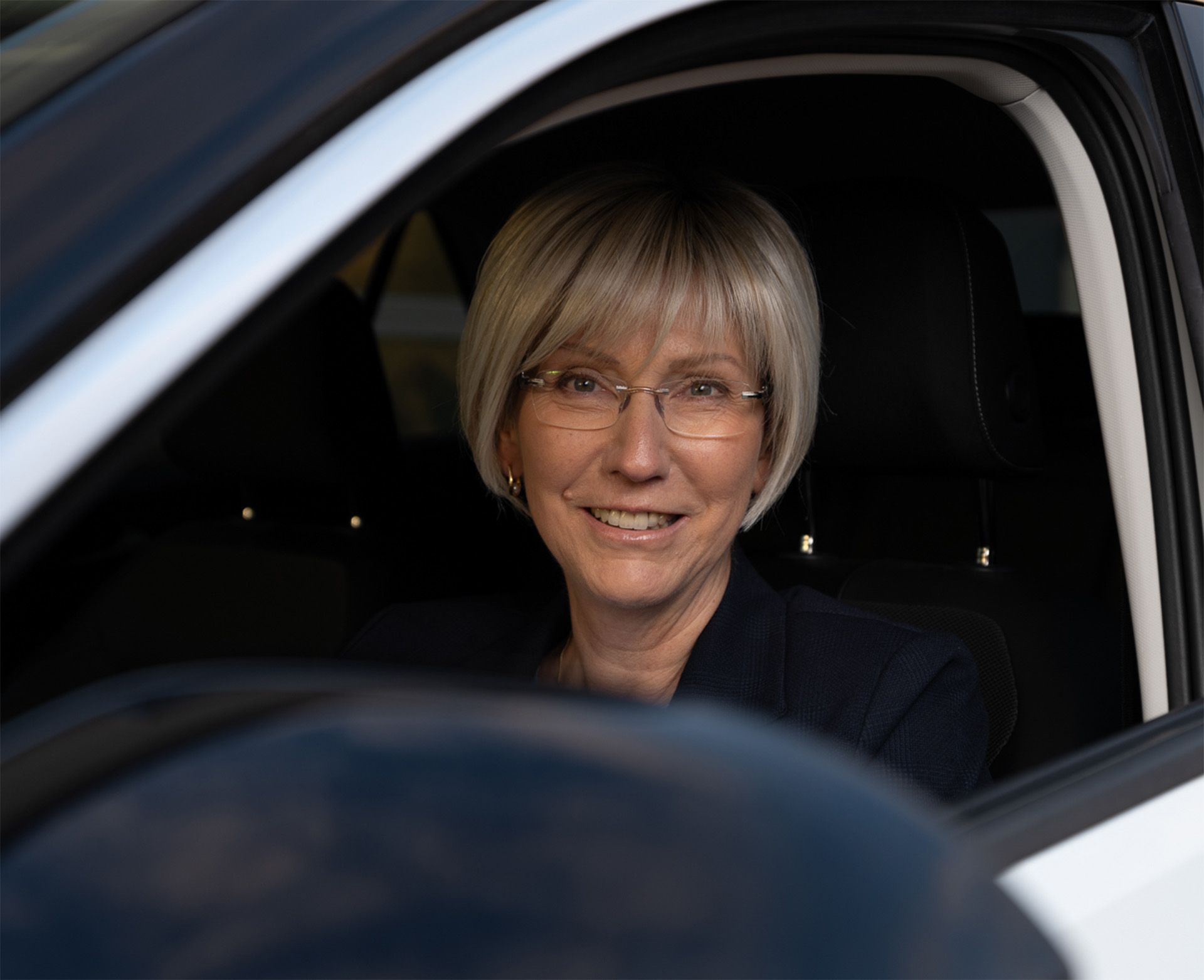 Eine professionell gekleidete Frau im Auto lächelt – repräsentativ für die familiengeführte Peter Kröner GmbH, die Injektoren und Zubehör für die Partikelschaumindustrie entwickelt und fertigt.