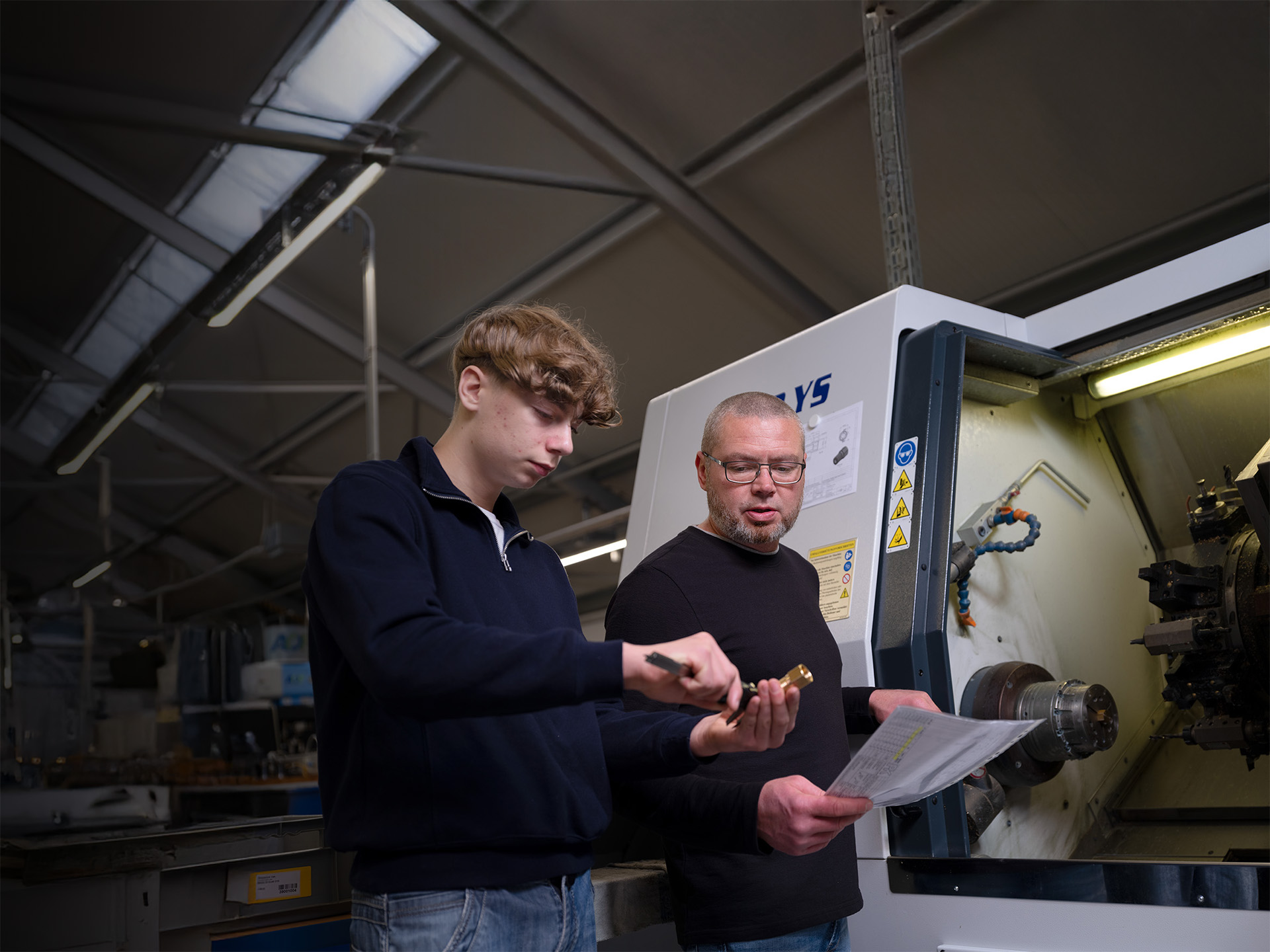 Zwei Mitarbeiter in der Werkstatt von Peter Kröner GmbH, einem familiengeführten Unternehmen für Partikelschaumindustrie-Zubehör, prüfen ein Bauteil und ein Dokument an einer CNC-Maschine – Abbildung der präzisen Fertigung und Ausbildung im Unternehmen.