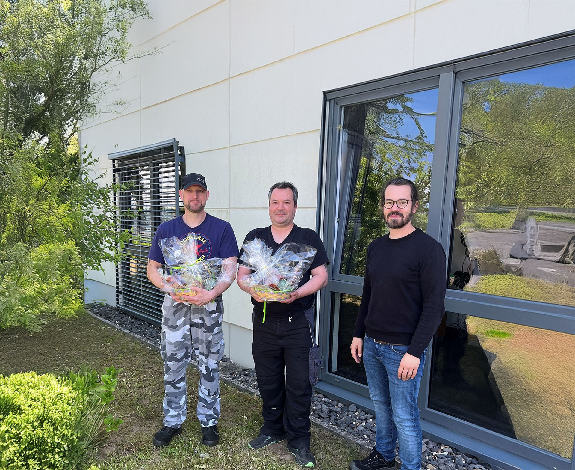 Drei Mitarbeiter der Peter Kröner GmbH, einem familiengeführten Stahlunternehmen, stehen vor einem modernen Firmengebäude mit großen Fenstern und Grünanlagen; zwei halten verpackte Geschenkkörbe, vermutlich bei einem internen Event oder Anerkennungsakt.