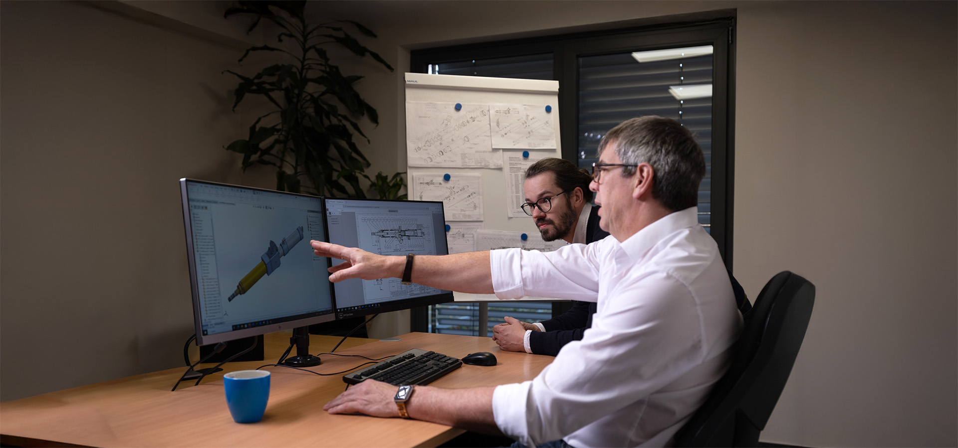 Zwei Mitarbeiter der Peter Kröner GmbH betrachten am Arbeitsplatz ein 3D-Modell eines Injektors auf zwei Monitoren, im Hintergrund ein Whiteboard mit technischen Zeichnungen und eine Pflanze.