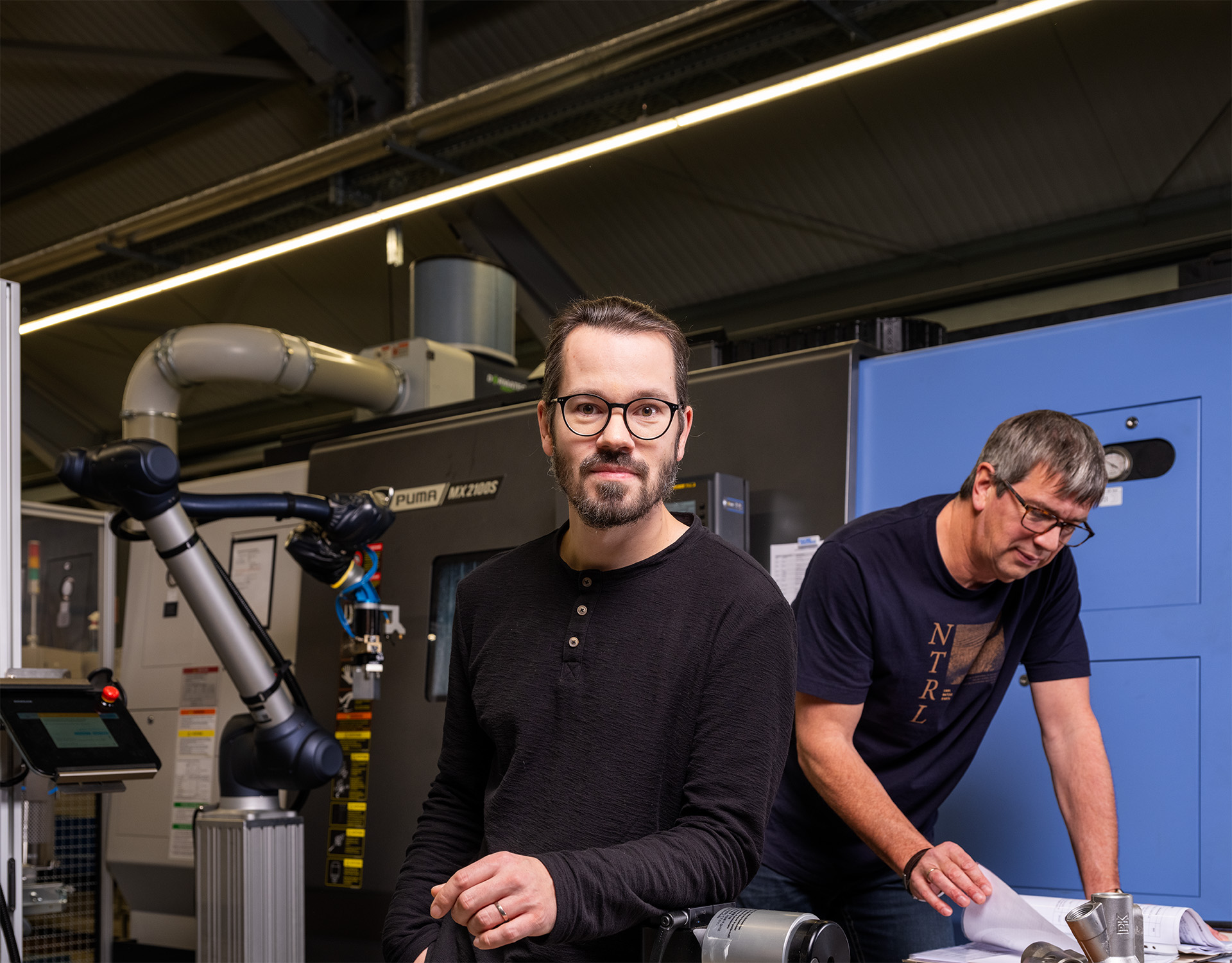 Zwei Mitarbeiter von Peter Kröner GmbH arbeiten in der Produktionsstätte mit industrieller Ausrüstung, darunter einem Roboterarm, für die Herstellung von Injektoren und Zubehör für die Partikelschaumindustrie.