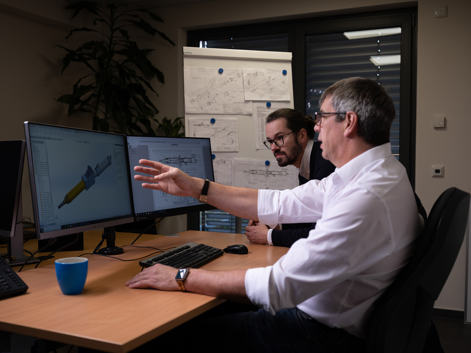 Zwei Mitarbeiter der Peter Kröner GmbH diskutieren an einem Schreibtisch mit zwei Monitoren technische Zeichnungen von Injektoren für die Partikelschaumindustrie.