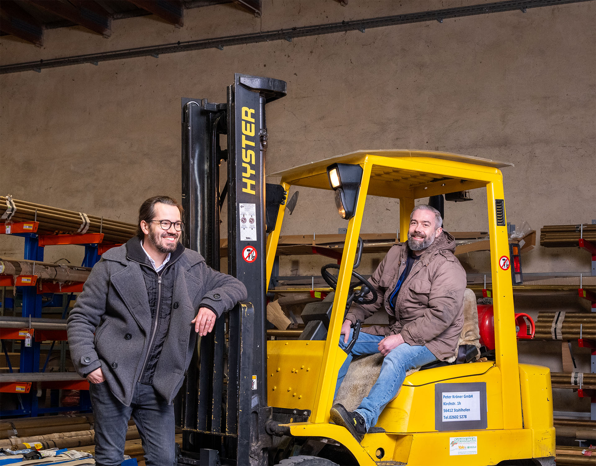 Zwei Mitarbeiter der Peter Kröner GmbH in einem Lager mit einem gelben Hyster Gabelstapler – das familiengeführte Unternehmen aus Stahlhofen fertigt Injektoren und Zubehör für die Partikelschaumindustrie.